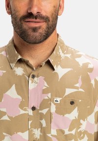 Kurzärmliges Hemd mit Knöpfen in Braun mit rosa-weißen Blumenmuster. Verfügt über eine Brusttasche und ein Logopatch auf der Tasche.