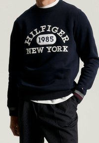 Mężczyzna w granatowym sweatshirtcie Hilfiger z białym haftem "1985 New York", w połączeniu z ciemnymi spodniami i białą koszulką pod spodem.