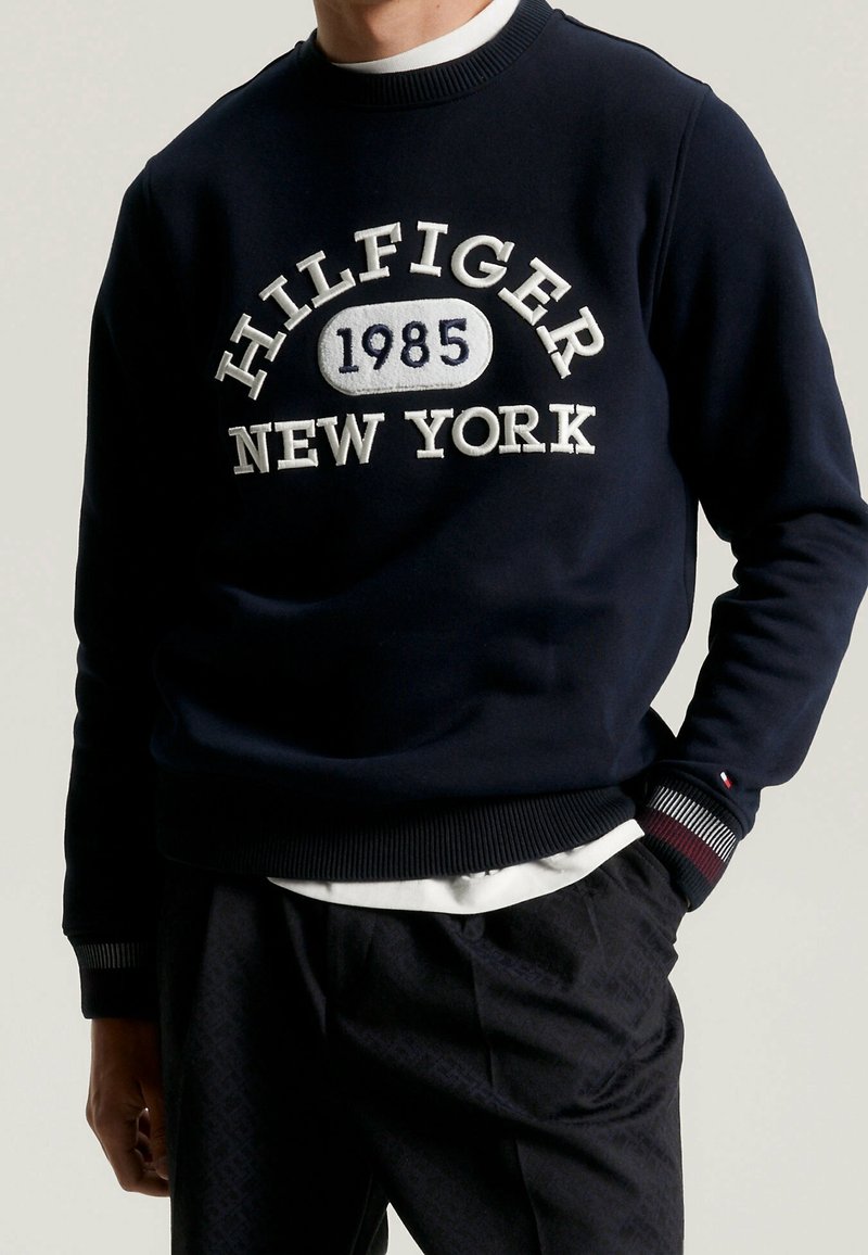 Mężczyzna w granatowym sweatshirtcie Hilfiger z białym haftem "1985 New York", w połączeniu z ciemnymi spodniami i białą koszulką pod spodem.