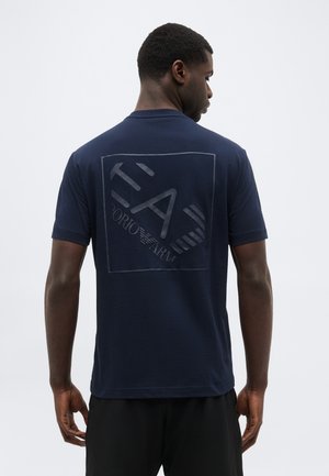 Egy férfi háttal áll, sötétkék pólót visel, amelynek hátoldalán nagy Emporio Armani logó látható, egyszínű fehér háttér előtt.