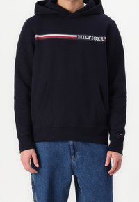 Felpa blu navy con tasca a marsupio frontale, design decorativo a righe rosse e bianche, e logo "HILFIGER" ricamato sul petto.