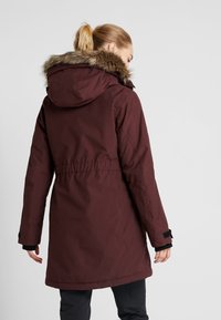 Burgunderfärgad parkas med en huva som är fodrad med konstpäls, elastisk midja och svarta detaljer. Har ett vattentåligt material och en medellång design.