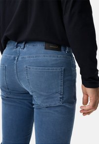 Blaue Cordjeans mit schmaler Passform, fünf Taschen, sichtbaren Nähten und einem schwarzen Lederetikett am Bund.