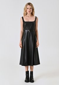 Schwarzes Kunstlederkleid mit quadratischem Ausschnitt, verstellbarem Gürtel in der Taille und einem ausgestellten Rock. Kombiniert mit klobigen schwarzen Knöchelstiefeln.