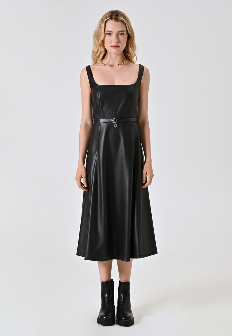 Schwarzes Kunstlederkleid mit quadratischem Ausschnitt, verstellbarem Gürtel in der Taille und einem ausgestellten Rock. Kombiniert mit klobigen schwarzen Knöchelstiefeln.