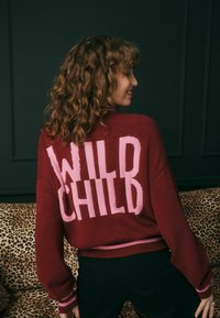 Raudona megztinė su užrašu "WILD CHILD" rožine spalva ant nugaros, su raukšlėmis ant rankovių ir apatinio krašto bei kontrastinga rožine juostele.
