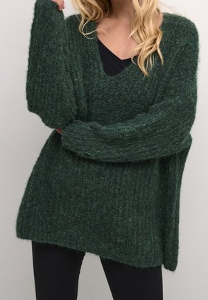 Femme portant un pull en maille duveteux vert foncé surdimensionné avec un col en V profond par-dessus un haut noir, associé à un pantalon noir, posant les bras croisés.