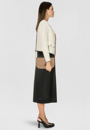 Beige schoudertas van glad leer, met een klep met sluiting en subtiel logo, gedragen bij een getextureerd wit jasje en een zwarte rok.