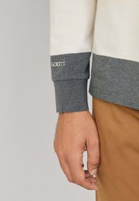 Ilga rankovėmis marškiniai su kreminių, pilkų ir tamsiai pilkų horizontalių dryžių raštu. Yra pilka rankogalių detalė su siuvinėjamu "HACKETT" logotipu. Pagaminta iš medvilnės.