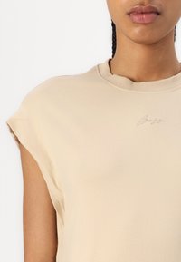 T-shirt beige à manches courtes en tissu doux avec une texture subtile ; il présente un logo cousu sur la poitrine et un col rond.