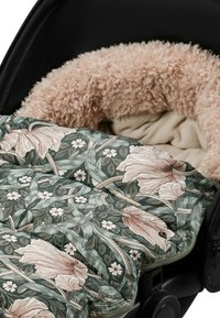 Beigen väriset kiharat turkkiset koira lepää kukallisella vihreällä ja vaaleanpunaisella pehmustetulla peitolla mustassa lemmikkivuoteessa tai -kantokopassa.