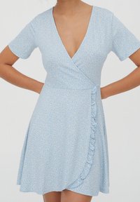 Vestido envolvente azul claro con mangas cortas, que presenta un patrón floral, escote en V y un detalle de volantes en la parte delantera. Tela suave y texturizada.