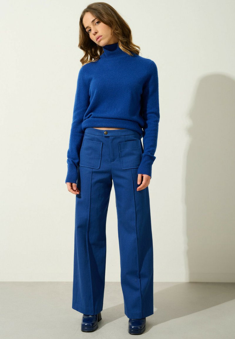 Blauer Wollrollkragenpullover mit langen Ärmeln, kombiniert mit hochgeschnittenen blauen Hosen mit aufgesetzten Taschen und einem vorderen Knopfverschluss.