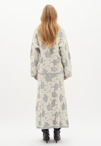 Donna con lunghi capelli biondi, che indossa un maglione floreale grigio a trama e una gonna lunga coordinata, con stivali neri con tacco, in piedi di spalle.