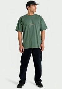 Green cotton t-shirt with a loose fit and short sleeves, featuring a small embroidered logo on the chest. Paired with dark denim pants.
