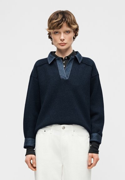 Jeune personne aux cheveux courts et bouclés portant un pull marine avec col et poignets en jean, une chemise noire en dessous et un pantalon blanc, debout immobile.