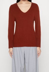 Pullover ruggine a V con maniche lunghe. Realizzato in tessuto a maglia testurizzata. Presenta una vestibilità rilassata e dettagli a costine sui bordi.