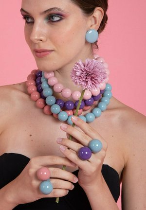 Donna che indossa gioielli in perline spesse dai colori pastello, mentre tiene un fiore di crisantemo rosa, su uno sfondo rosa.