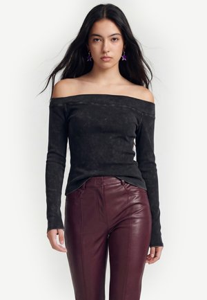 Kvinde med langt sort hår har en sort top med bare skuldre, kastanjebrune læderbukser og lilla stjerneformede øreringe mod en hvid baggrund.