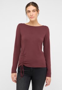 Ilgos rankovės bordo spalvos palaidinė su apvalia iškirpte, šoniniu raukiniu ir raišteliu apačioje. Lygi audinio tekstūra. Derinama su juodais džinsais.