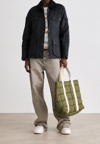 Černá prošívaná bunda s předními kapsami, kombinovaná s pruhovanou košilí a béžovými kalhotami. Drží olivově zelenou polstrovanou tašku s krémovými popruhy.