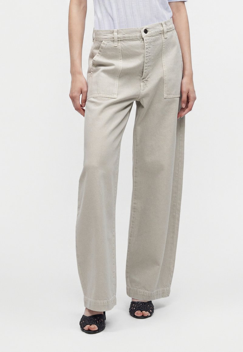 Femme portant un pantalon large beige clair avec des poches plaquées à l'avant et des sandales tissées noires à bout ouvert, debout devant un fond uni.