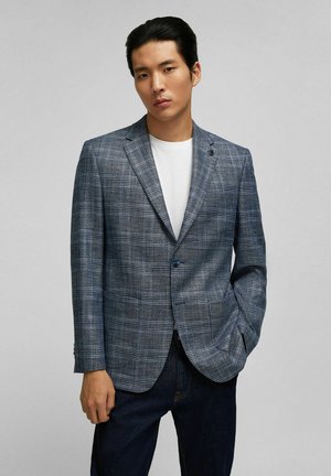 Blazer in einem blauen Karomuster, aus strukturiertem Stoff. Verfügt über einen einzigen Knopf, einen Reverskragen und zwei Fronttaschen. Getragen über einem weißen Hemd.