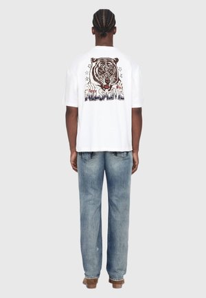 Man with braided hair seen from behind wearing a white oversized t-shirt with a tiger and "ALLSAINTS" graphic and faded blue jeans.
