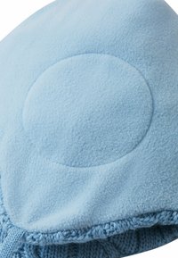 Coperta in pile di colore azzurro chiaro con una superficie liscia, forma arrotondata e bordo a maglia lungo il bordo inferiore. Presenta un design circolare prominente.
