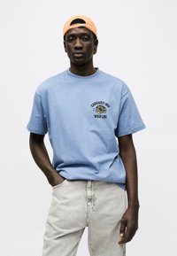 Νεαρός άνδρας που φοράει ένα ανοιχτό μπλε t-shirt Carhartt WIP Wild Life, ανοιχτό γκρι τζιν και πορτοκαλί καπέλο φοράει ανάποδα, στέκεται με το ένα χέρι στην τσέπη.