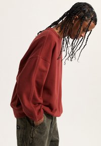 Sudadera marrón con hombros caídos, fabricada en algodón texturizado. Lleva puesta sobre pantalones de mezclilla verdes con un bolsillo lateral visible y un parche con el logo.