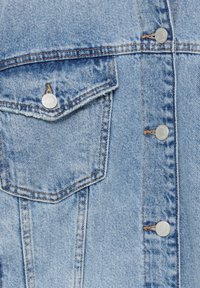 Vaaleansininen farkkutakki, jossa on terävä tasku, hopeiset nepparit ja kontrastivärinen ommel. Kankaan tekstuuri on klassinen.