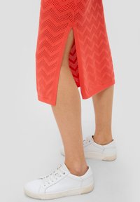 Koraalrood perforated jurkje met een zigzagpatroon, voorzien van zijsplitten. Gepaard met witte sneakers, wat een moderne casual uitstraling benadrukt.