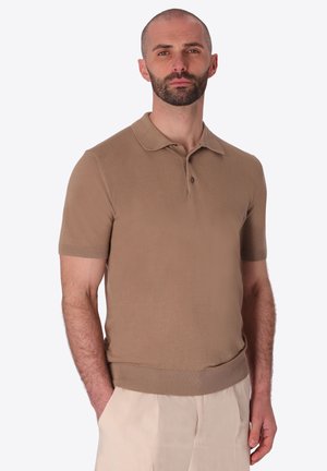 Uomo con barba e testa rasata che indossa una polo marrone a maniche corte e pantaloni beige chiaro, in piedi con una mano in tasca.