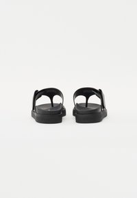 Sorte sandaler med minimalistisk design, som har en båndrem, teksturert fotseng og solid gummisåle, sett fra baksiden.