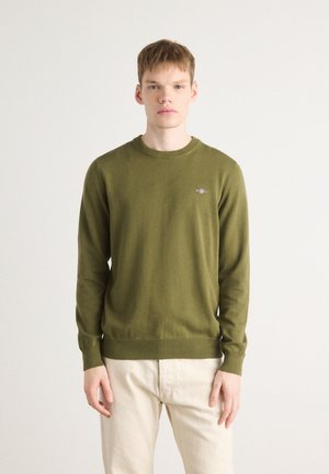 Pull olive en tissu doux. Col rond, manches longues et poignets côtelés. Petit logo brodé sur la poitrine. Associé à un pantalon clair.