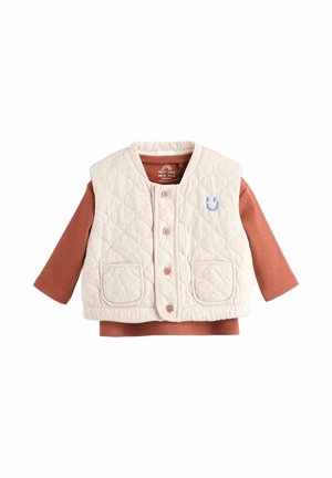 Bežs šūnu zīdaiņu veste ar pogām virs rūsas krāsas garām piedurknēm ribota krekla, uz kura krūšu daļas ir maza zila smaidoša seja.