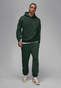 Ciemnozielona bluza z kapturem z przednią kieszenią, sznurkami, dopasowane dresy oraz beżowe sneakersy. Casualowy, luźny krój i gładka faktura.