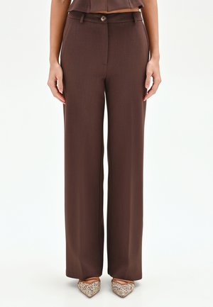 Donna che indossa pantaloni marroni a vita alta e gamba larga con bottoni frontali e ballerine intrecciate con punta appuntita, in piedi su uno sfondo bianco uniforme.