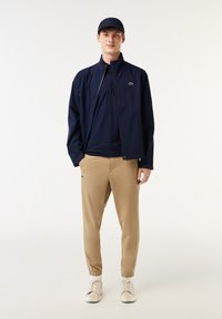 Marine lichtgewicht jack, gestreepte marine polo, beige tapered broek met cuffs, witte sneakers en een marine cap met logo accent.