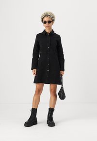 Crna traper haljina s ovratnikom i prednjim kopčanjem na gumbe. Uparena s crnim čizmama na vezanje i malom torbom.