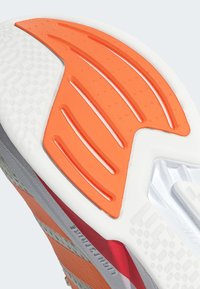 La suela del zapato presenta un diseño texturizado en gris y blanco, con un acento en naranja brillante y ranuras para tracción, mostrado desde una perspectiva en ángulo.