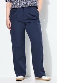 Persona che indossa pantaloni larghi blu navy con un sottile motivo a pois, una camicia a motivi parzialmente inserita e sneaker bianche con suola color gomma.