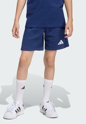 Dziecko ubrane w granatowe szorty i bluzkę, białe skarpetki Adidas oraz białe buty sportowe z czarnymi paskami, stoi na jednolitym tle.