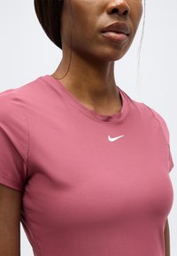 Γυναίκα που φοράει ροζ εφαρμοστή μπλούζα Nike, ορατή από το λαιμό έως το στήθος, με το λευκό λογότυπο Nike swoosh μπροστά.