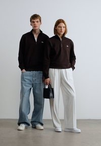 Καφέ πουλόβερ με φερμουάρ, ανοιχτό μπλε φαρδιά τζιν και λευκά αθλητικά σε ένα μοντέλο· λευκό πλισέ παντελόνι και μαύρη τσάντα στο άλλο.