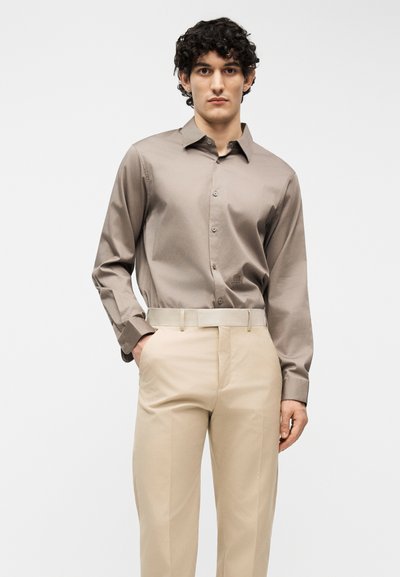 Άνδρας με σγουρά σκούρα μαλλιά φοράει πουκάμισο σε χρώμα ταουπέ με μακριά μανίκια και κουμπιά, το οποίο είναι μπουσουλιασμένο σε μπεζ ψηλόμεσα παντελόνια με ταιριαστή ζώνη.