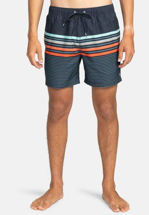 Man in een donker zwembroek met horizontale strepen in lichtblauw, wit en oranje, staand op blote voeten tegen een witte achtergrond.