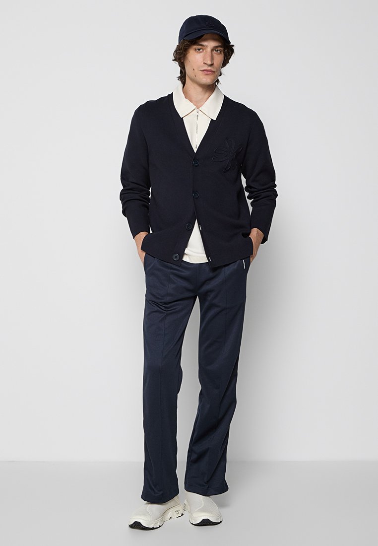 Marineblauer Cardigan mit Knopfverschluss und floralem Stickerei, kombiniert mit einem weißen Hemd mit Kragen und marineblauen Hosen, abgerundet mit weißen Sneakers und einer Mütze.