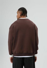 Bruine sweatshirt met een comfortabele pasvorm, oversized model, geribde zoom en boorden, met een witte binnenkraag, gedragen over een donkere spijkerbroek.